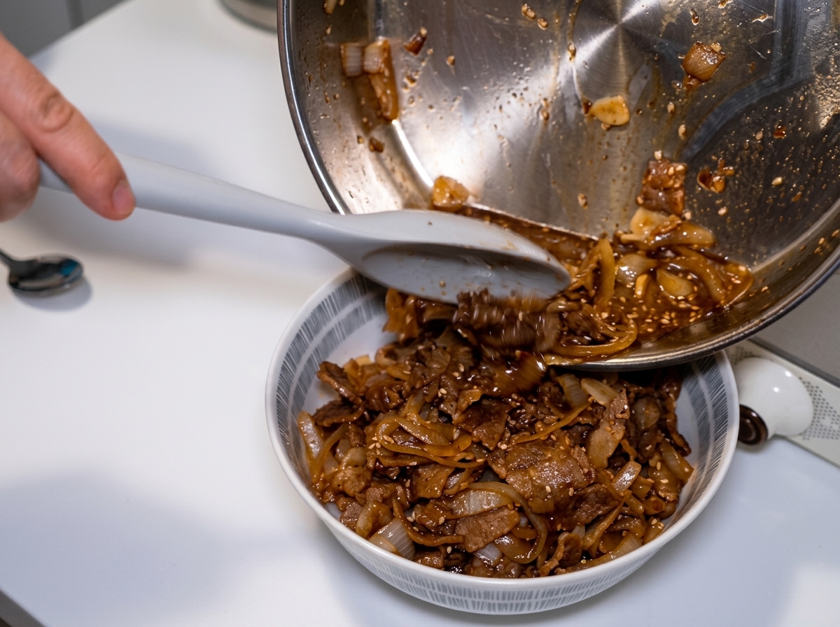 フライパンを傾け、スパチュラを使いながら、残った玉ねぎと旨味たっぷりのつゆを牛丼の上へと注いでいる様子。