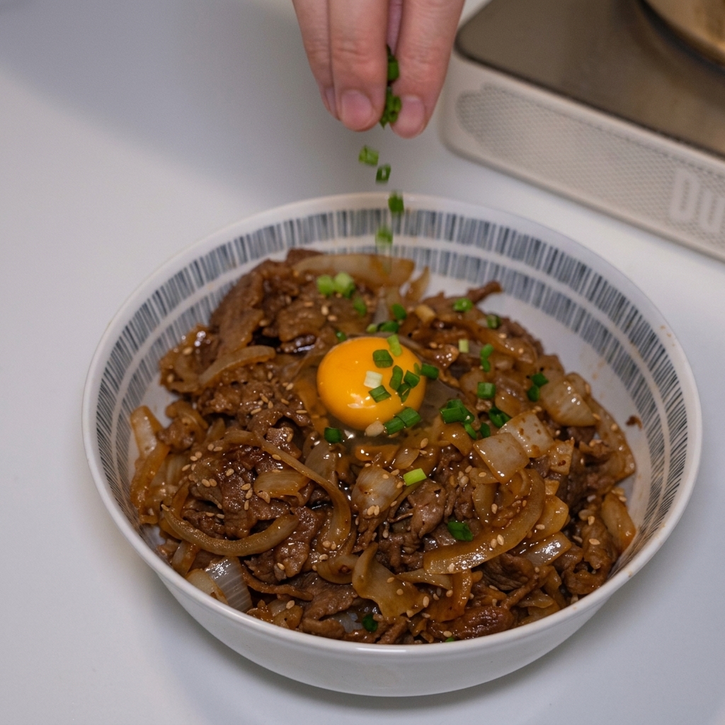 完成した卵黄付きの牛丼の上に、指先で刻んだ鮮やかな緑色の青ねぎを散らしている様子。