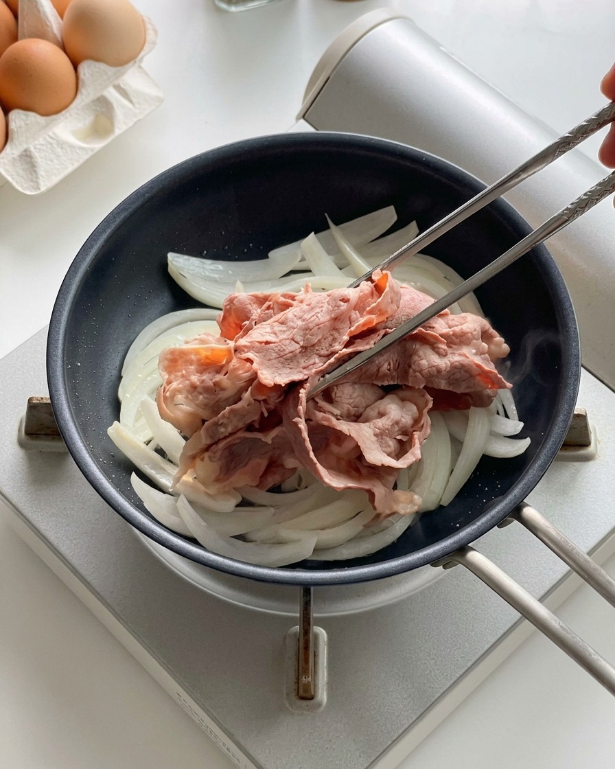 黒いフライパンで炒めている玉ねぎの上に、菜箸で湯通しした牛肉をのせている様子。