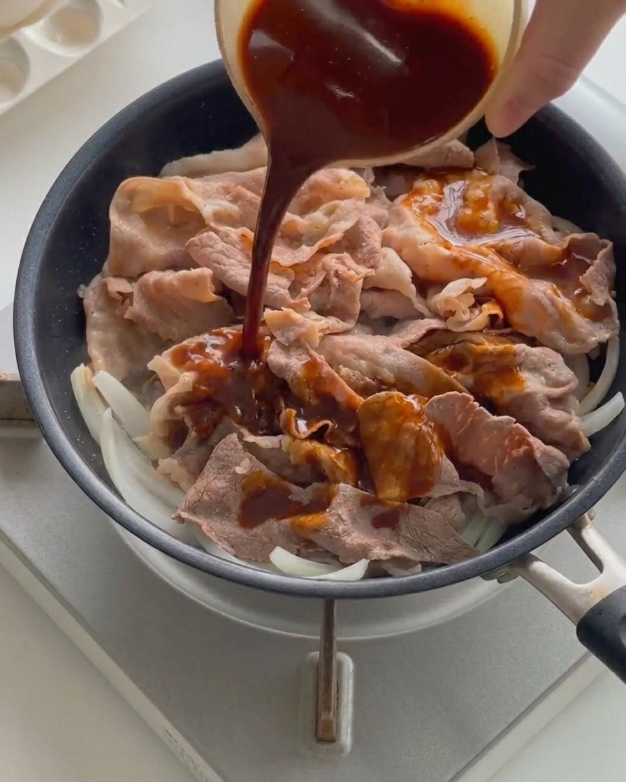 熱したフライパンの中の牛肉と玉ねぎの上から、小さなボウルに入った濃い茶色のタレを回し入れている様子。