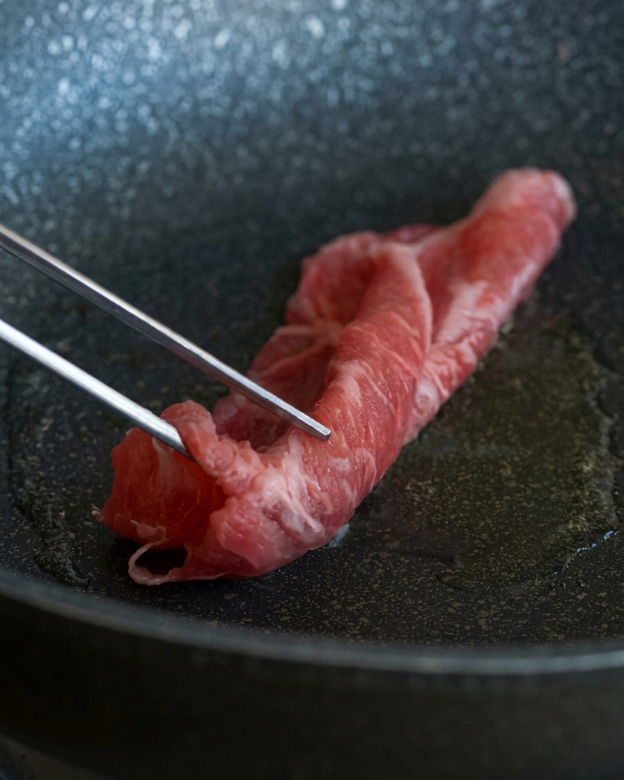 油がなじんだ熱いフライパンに、金属製のトングで霜降りの牛薄切り肉を入れる様子
