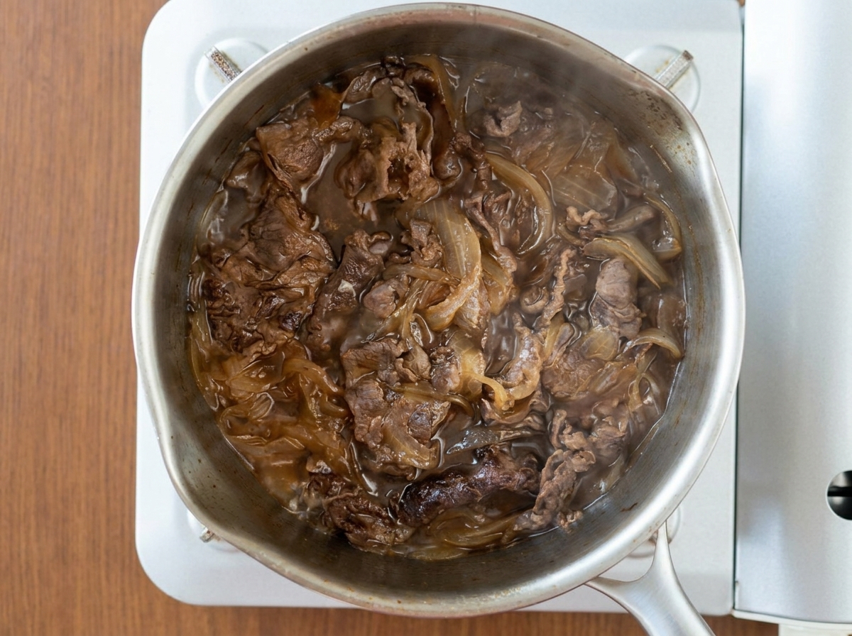 ステンレス鍋の中で、濃いめのタレと一緒に牛肉と玉ねぎがコトコトと煮えている様子の俯瞰
