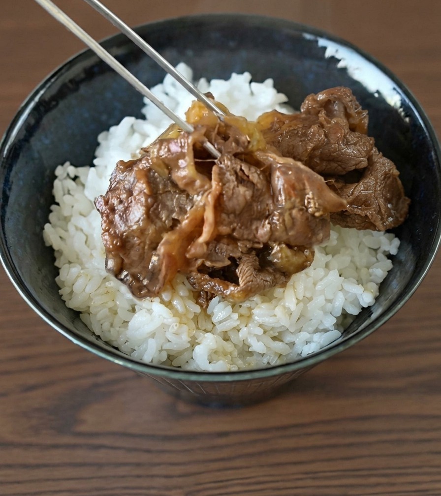 シックな陶器の器に盛られたふっくらとした白米の上に、金属製の箸で牛薄切り肉と玉ねぎをのせる様子