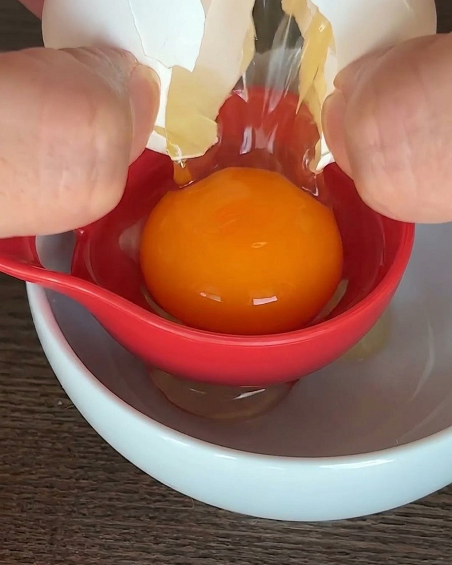 白いボウルの上で赤いエッグセパレーターを使い、中央に鮮やかなオレンジ色の卵黄だけを残す様子