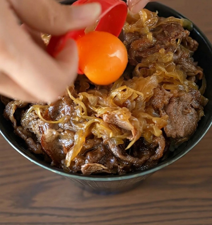 完成した牛丼の中央に、赤いセパレーターから鮮やかなオレンジ色の卵黄をそっと落とす様子
