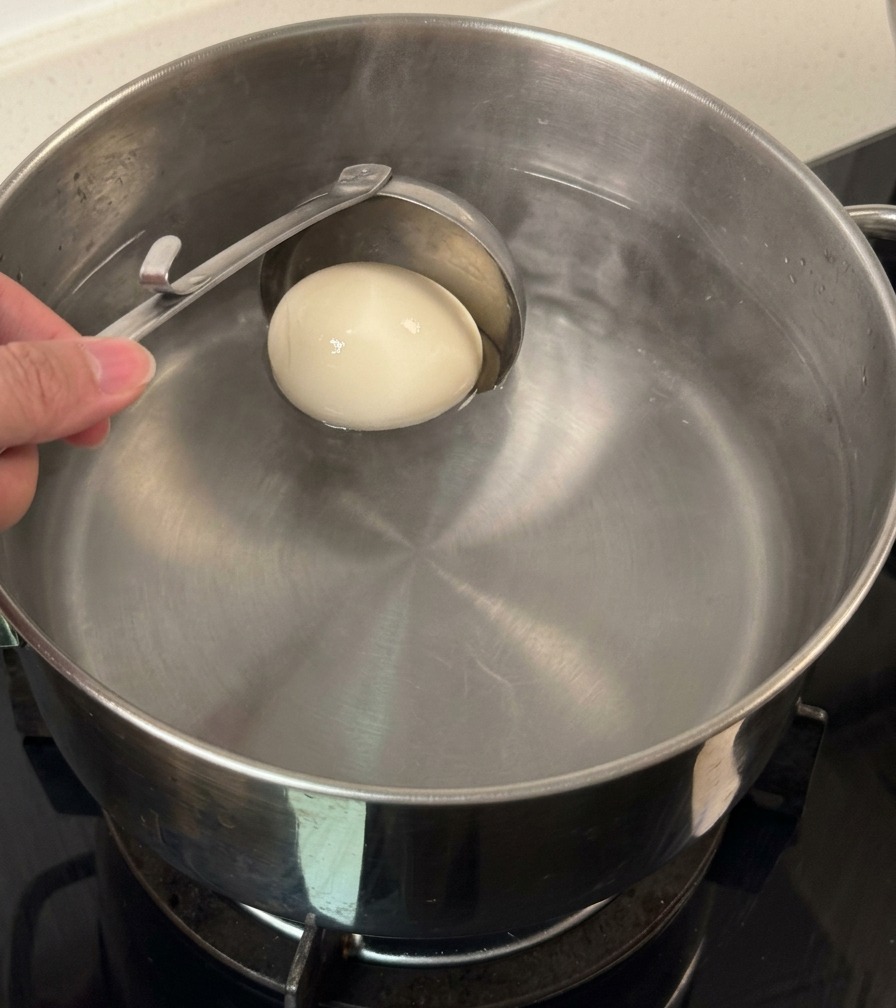 お玉を使って、殻付きの白い卵を1個、温度調整したお湯の入った鍋にそっと沈めている様子。
