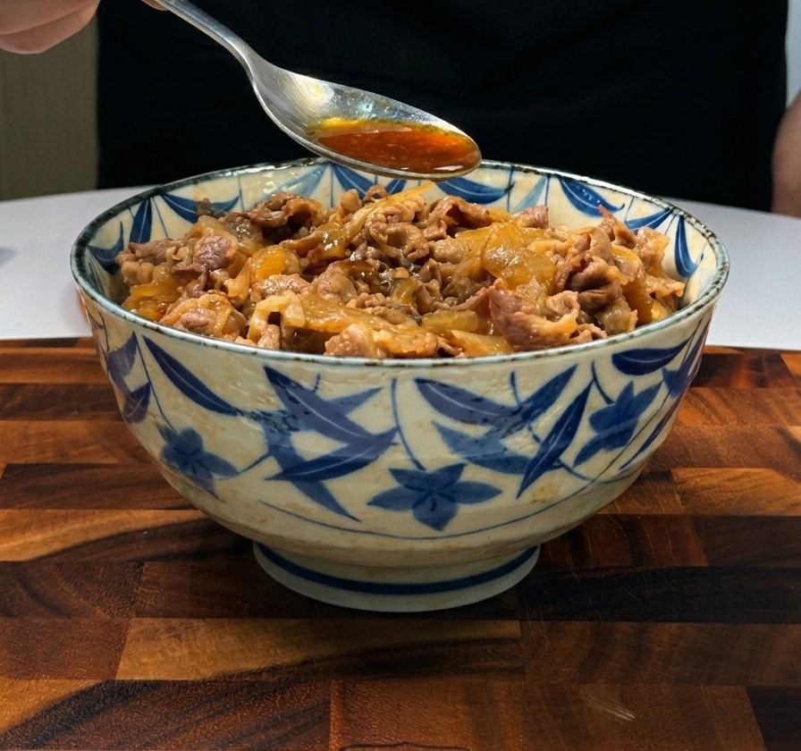 白いご飯の上に盛り付けられた薄切りの牛肉と玉ねぎの上から、大きめのスプーンで濃い茶色のつゆを回しかけている様子。