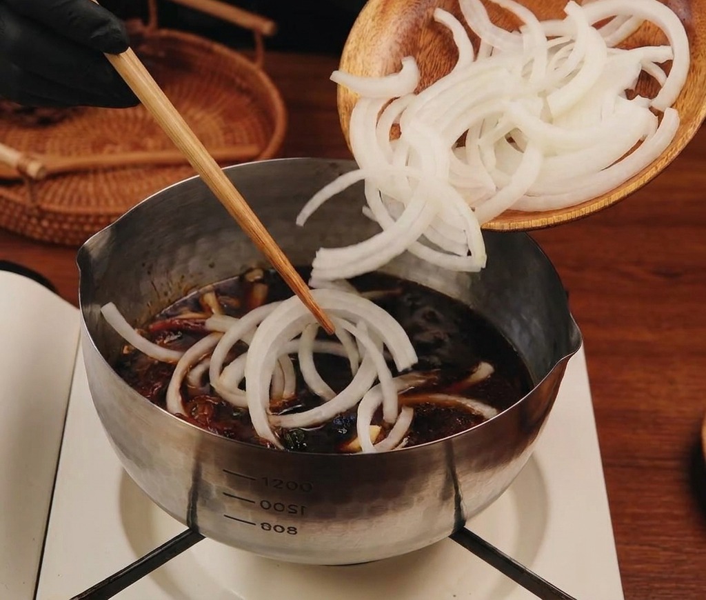 木製の皿から、薄切りにした白い玉ねぎが濃い色のタレが入ったステンレス鍋に投入されている様子。