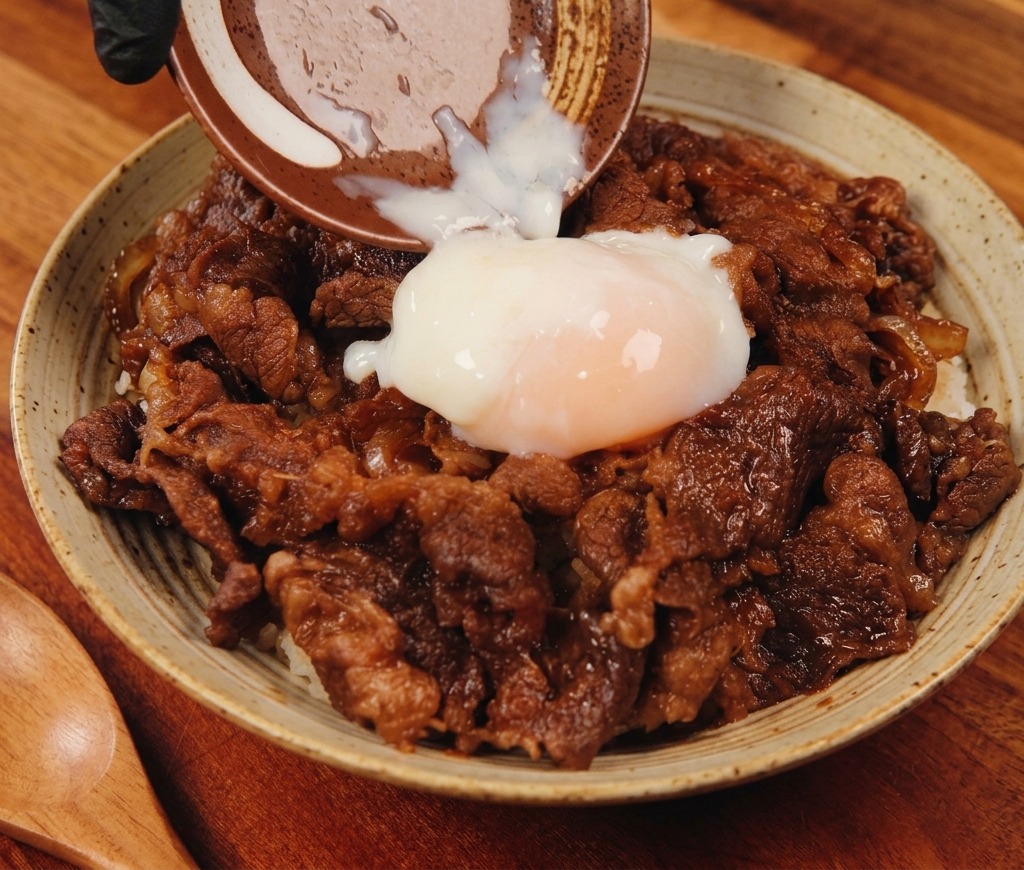 湯気が立つ美味しそうな牛丼の中央に、小さなセラミックの小皿からとろっとした温泉卵が優しく落とされている様子。