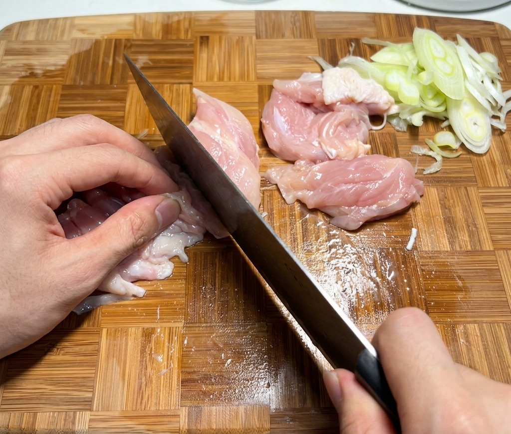 木製のまな板の上で、生の鶏もも肉を一口大の大きさに切っている様子。