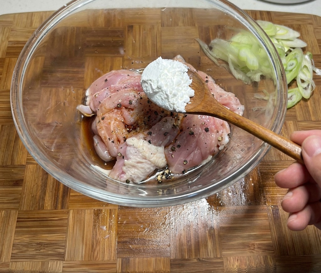 醤油やスパイスで下味をつけた生の鶏肉が入ったガラスボウルに、片栗粉をスプーン1杯加えている様子。