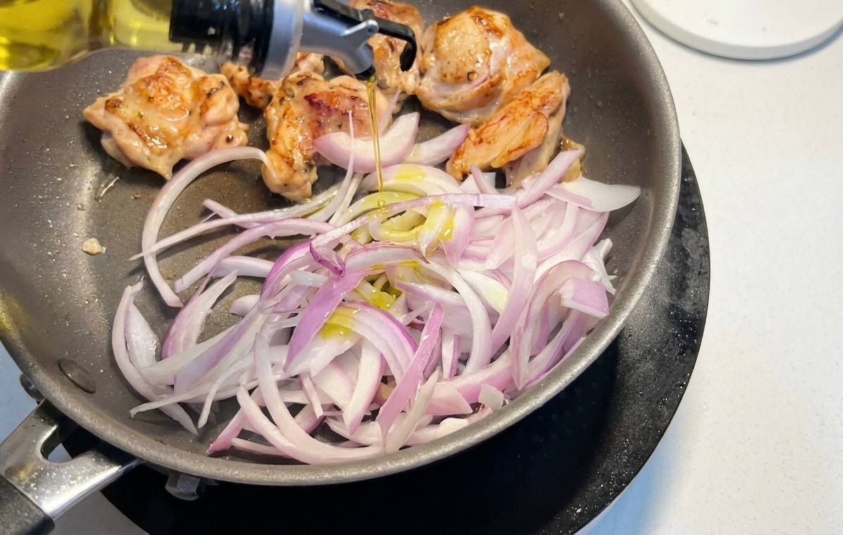 焼き色がついた鶏もも肉が入ったフライパンにスライスした赤玉ねぎを加え、上から油を回しかけている様子。