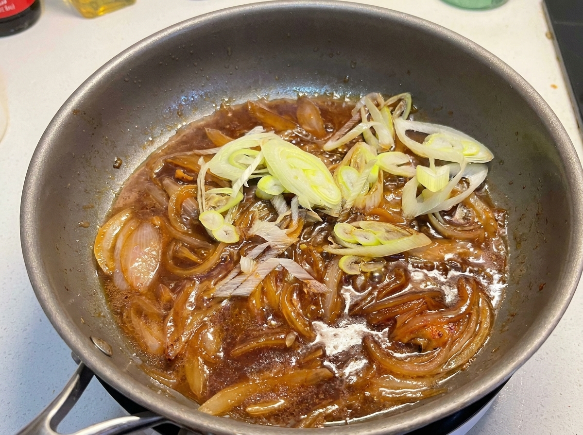 煮立っているタレと炒めた玉ねぎが入ったフライパンに、カットした青ねぎを加えている様子。
