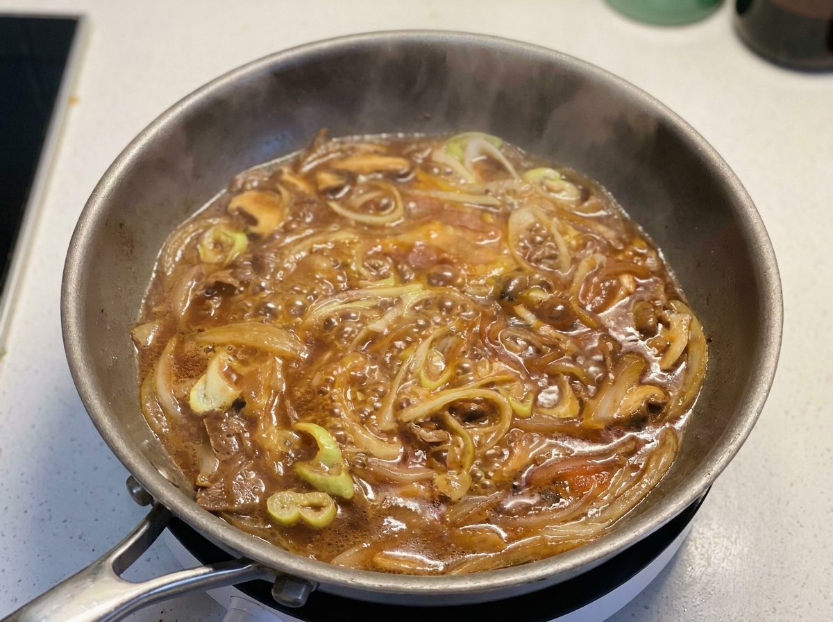 グツグツと煮立つタレ、青ねぎ、赤玉ねぎが入ったフライパンに、溶き卵を円を描くように回し入れている様子。