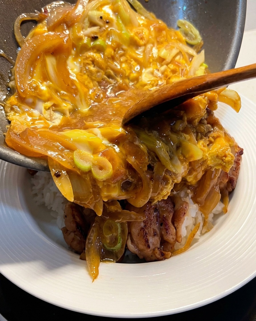 フライパンから、とろとろに火が通った卵と玉ねぎの具材を、あらかじめ鶏肉をのせておいた白米の丼の上にするりと滑らせている様子。