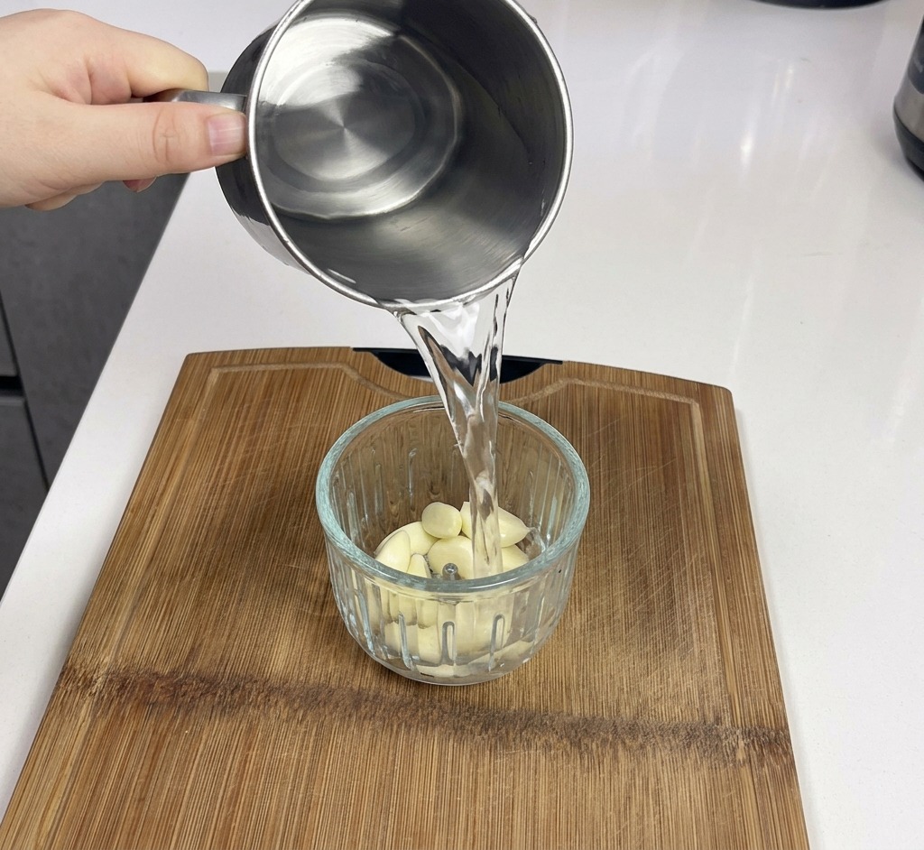 皮をむいた白い生ニンニクが詰まった透明なミキサーの容器に、金属製のマグカップから水を注いでいる様子。