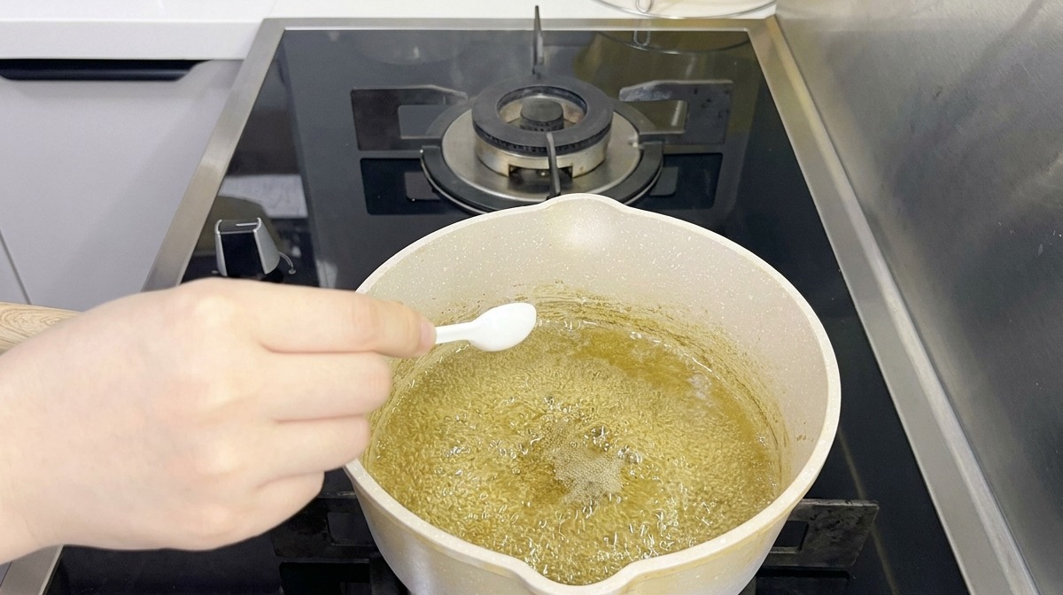 鍋の中で沸騰している少し緑がかった液体に、小さな白いスプーンを使って調味料を追加している様子。