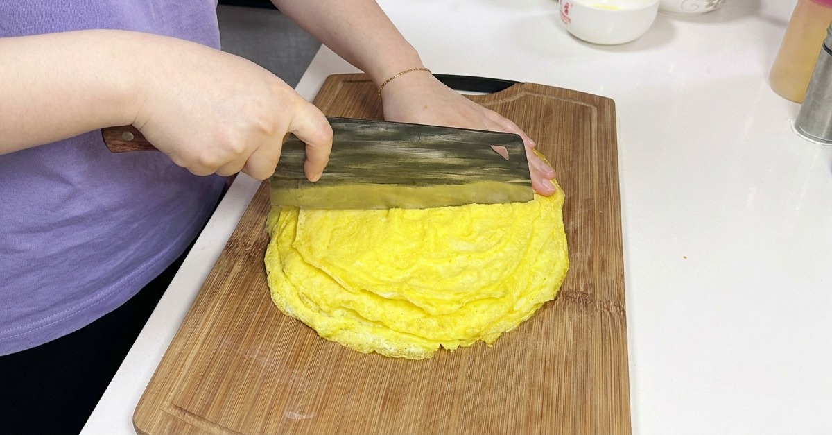 木製のまな板の上で、重ねられた薄く焼かれた黄色い卵のシートを、大きな中華包丁を使って切り分けている様子。
