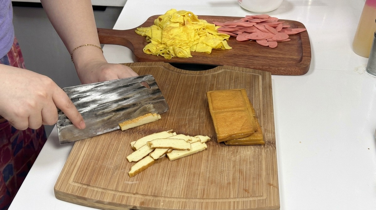 木製のまな板の上で、重ねられたきつね色の燻製豆腐のブロックを金属製の中華包丁で細切りにしており、背景には準備された卵とハムが映っている。