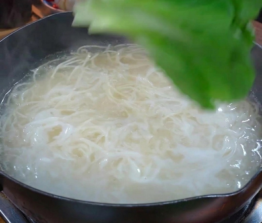 白い麺が茹でられている沸騰した鍋に、投入される新鮮な緑の青菜
