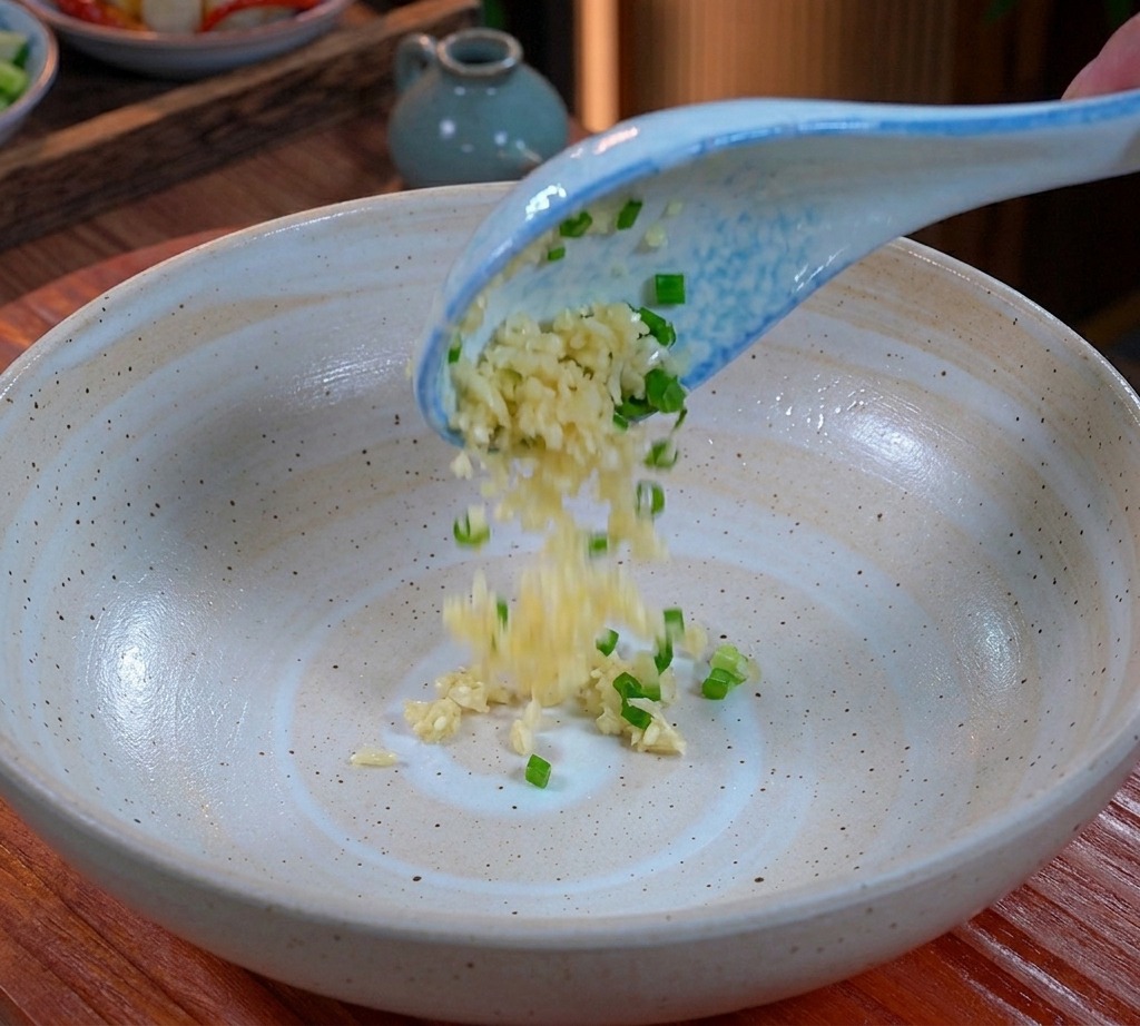 青と白のレンゲから、広口の磁器の器の底へと落とされるみじん切りのにんにくと刻みねねぎ