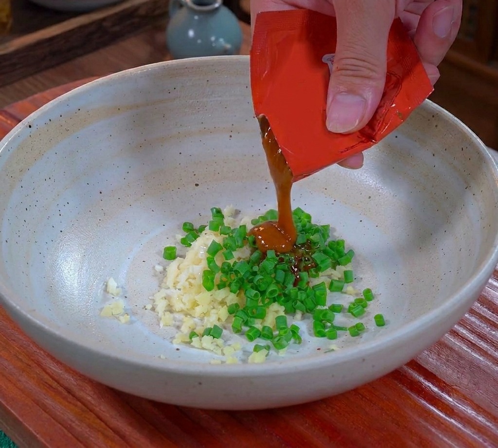 刻んだにんにくとねぎが入った磁器の器に、赤いパケットからとろみのある茶色の担々麺の素を絞り出す手