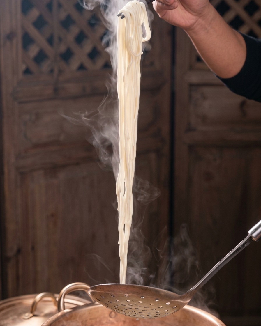 穴あきお玉と黒い箸を使って、湯気を立てる白い麺を銅鍋から持ち上げている様子。
