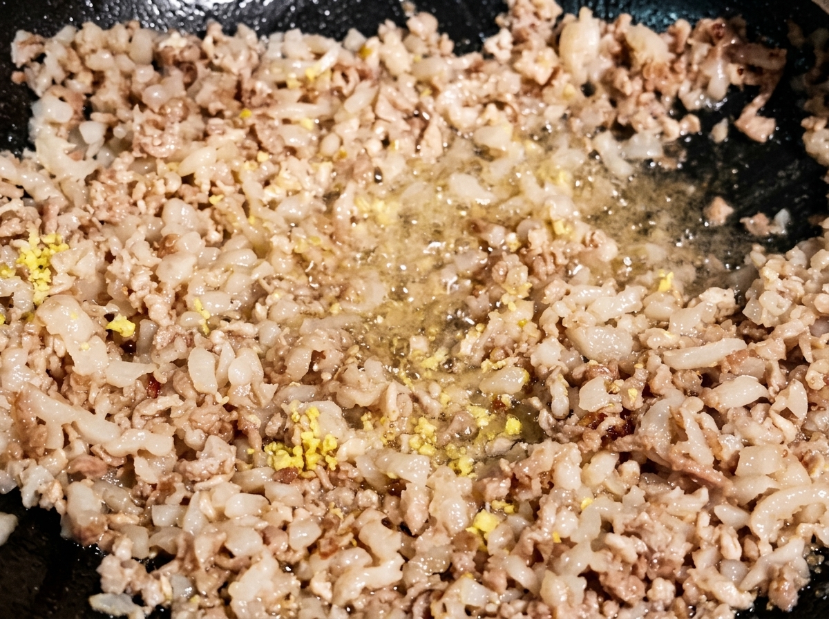 黒っぽい中華鍋の中で、ほんのり色づいた豚ひき肉にみじん切りの生姜とにんにくを加えて炒め合わせている様子。