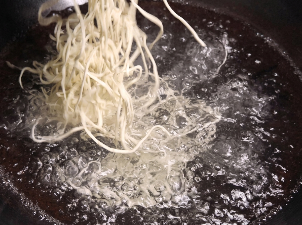 大きな鍋の中で激しく沸騰しているお湯の中に、生の白い中華麺の束を投入している様子。