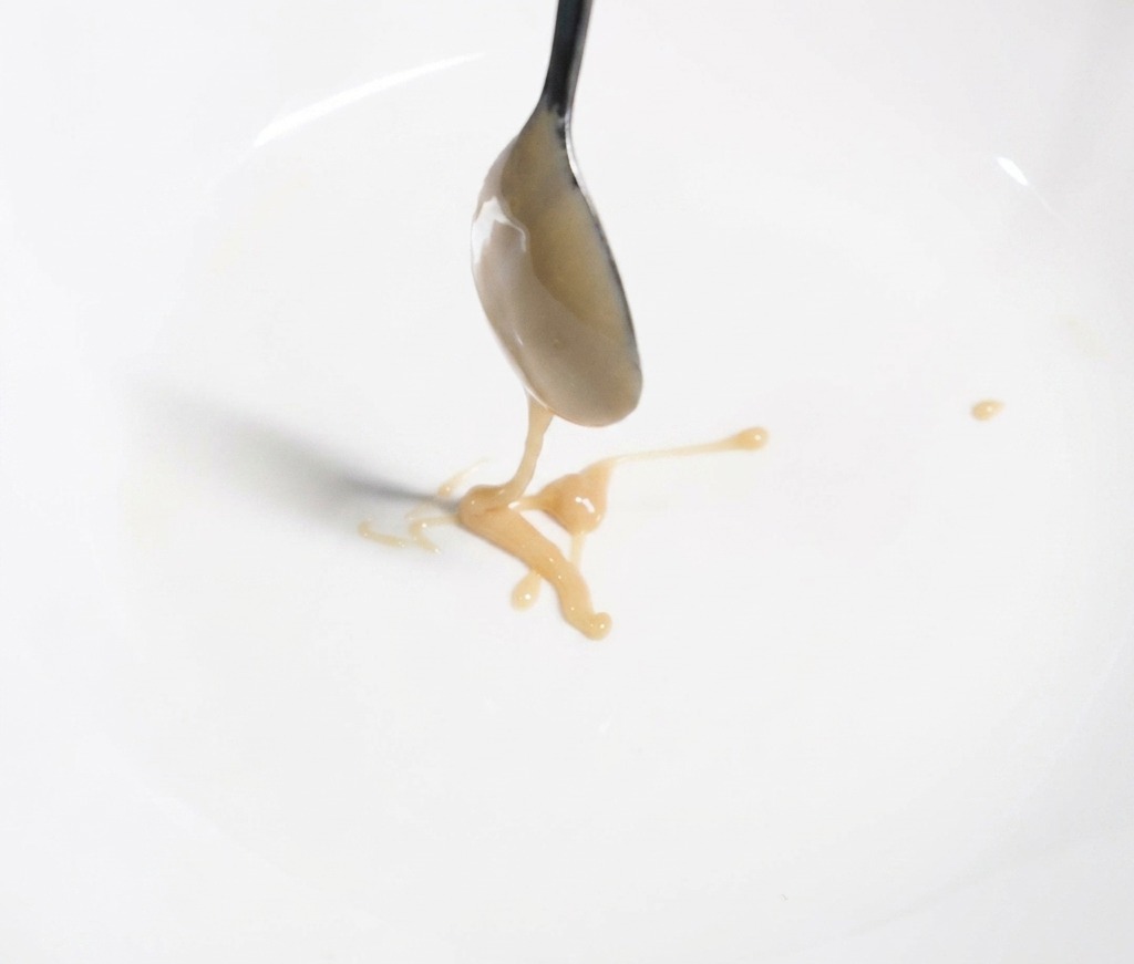 白い丼の中に、金属製のスプーンで濃厚な薄茶色の練り胡麻を落とし、麺のタレ作りの最初のステップを示している様子。