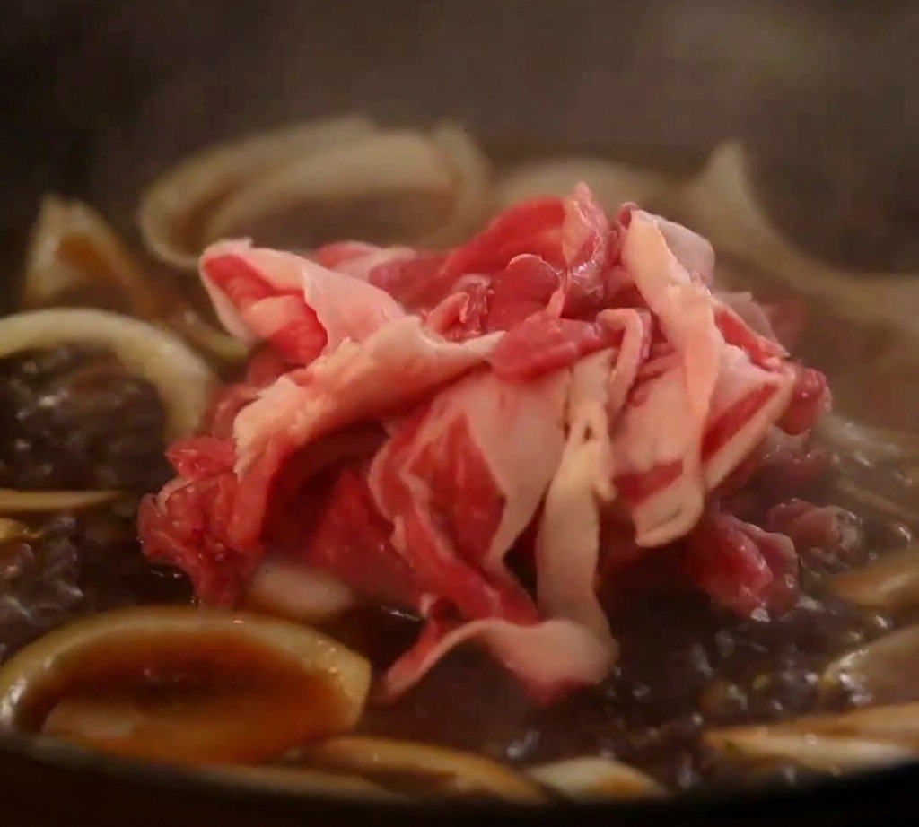 玉ねぎと和風のタレがぷつぷつと煮立つフライパンに、生の牛バラ肉のスライスを広げながら加えている様子。