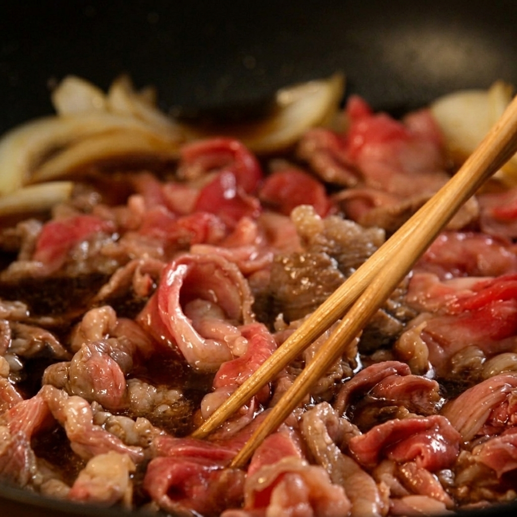 気泡が浮かぶ濃い茶色のタレの中で、薄切り牛肉と玉ねぎを木箸で優しく混ぜながら煮込んでいる様子。