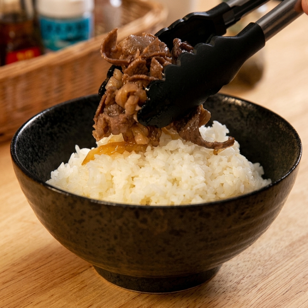 黒いトングを使い、炊きたての白いご飯を盛った丼の上に、煮上がった温かい牛肉と玉ねぎをたっぷりのせている様子。