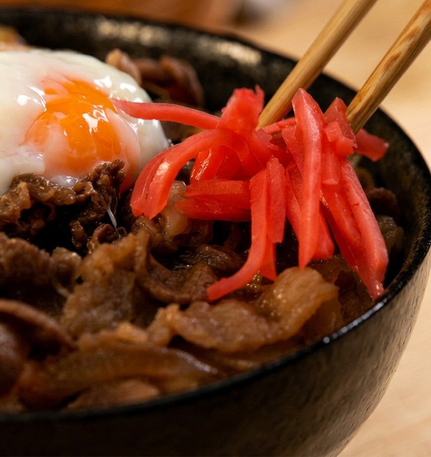 木箸を使い、温泉卵をのせた湯気立つ牛丼の脇に、鮮やかな赤色の紅生姜をそっと添えているクローズアップ写真。