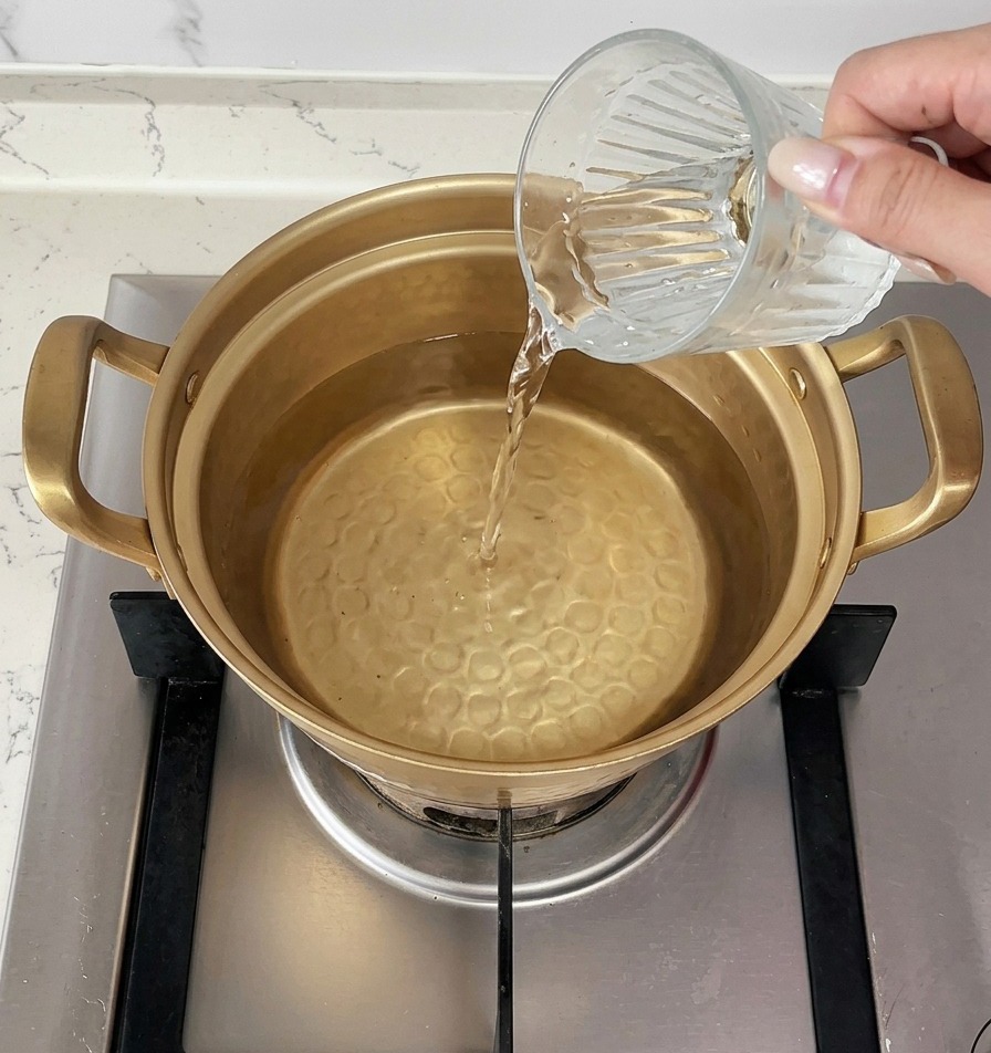 コンロの上で沸騰したお湯が入った金色の鍋に、片手で冷水を1カップ注ぎ入れている様子。