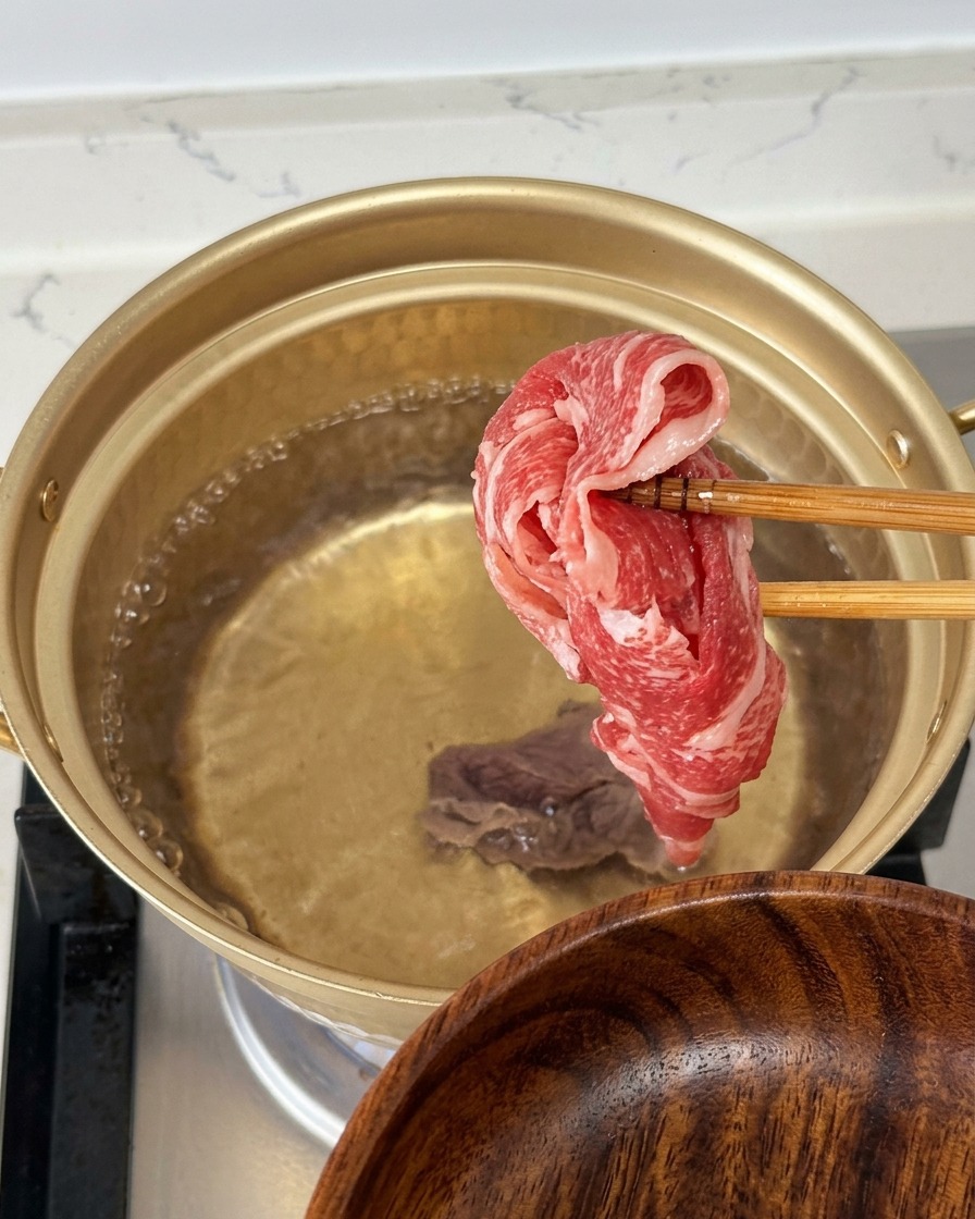 金色の鍋に沸騰したお湯の中で、箸を使ってきれいなサシの入った生の牛薄切り肉を1枚持ち上げている様子。