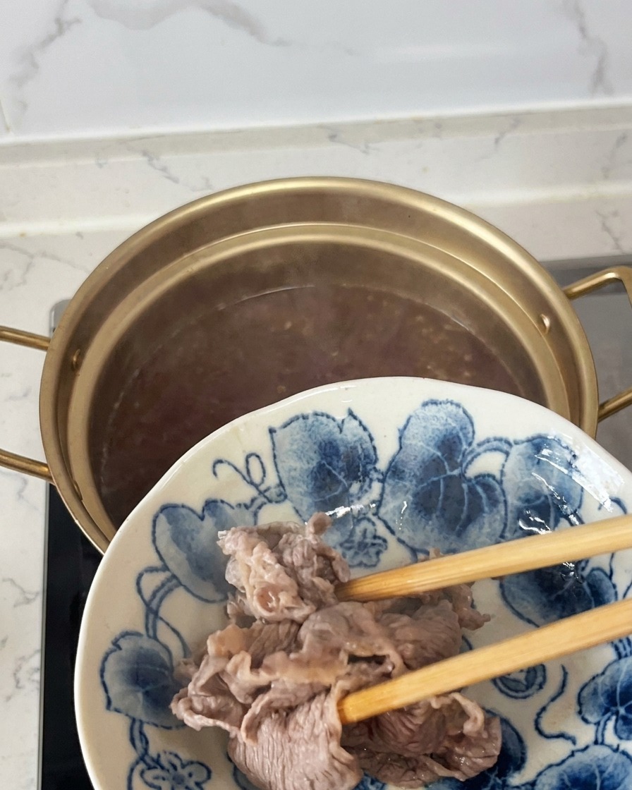 表面が白っぽく変化した半熟状態の牛肉を、箸で鍋から引き上げて青と白の柄の皿に移している様子。