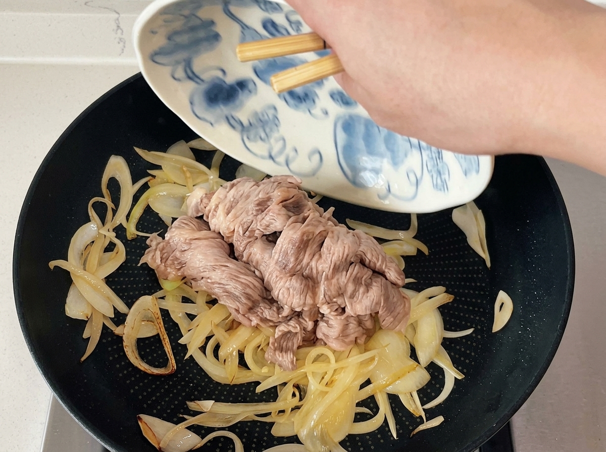 箸を使って、柄皿に用意しておいた湯通し済みの牛肉を、炒めた玉ねぎの入ったフライパンへ滑らせるように入れている様子。