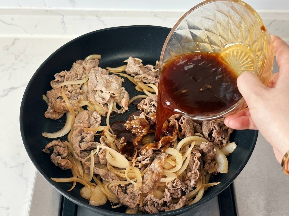 黒いフライパンの中の牛肉と玉ねぎの上へ、ガラスのボウルから濃い調味タレを一気に回し入れている様子。