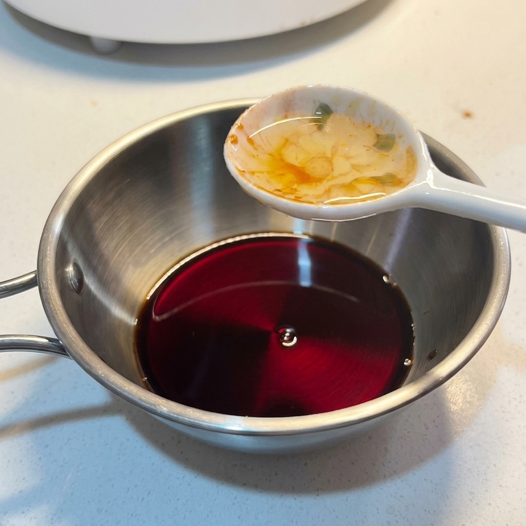 濃い色のタレが入った小さなステンレスボウルに、スプーンで液体調味料を加えている様子。