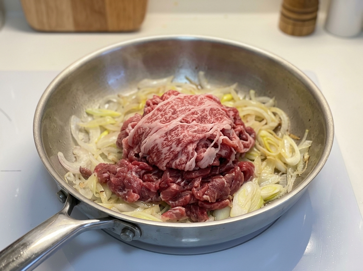 フライパンの炒めた玉ねぎの上に、生の牛薄切り肉をのせている様子。