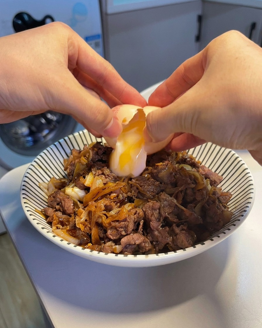 両手で温泉卵を割り、熱々の牛丼の中央に、とろりとした黄身と白身を落としている様子。