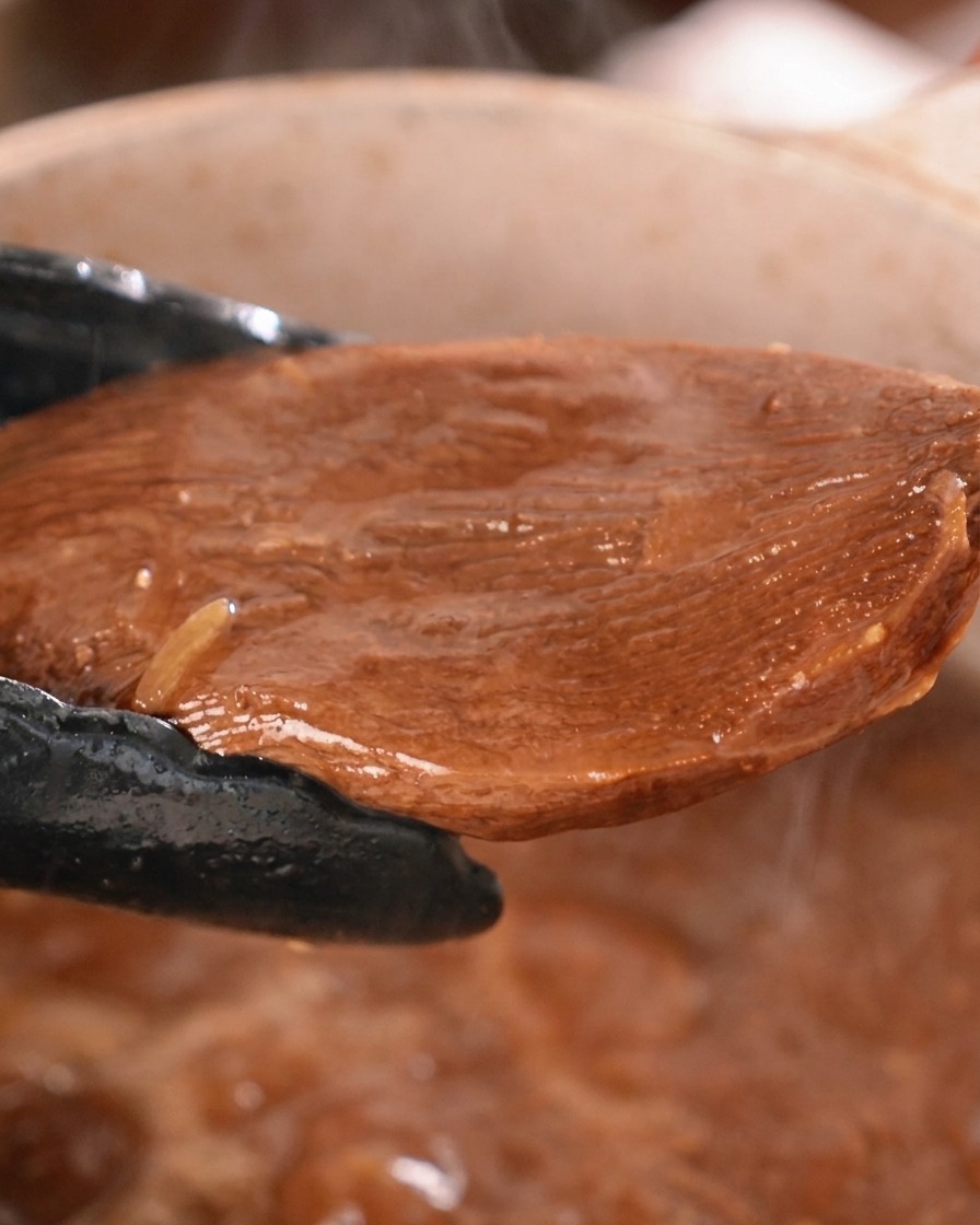 黒いキッチン用トングを使い、味がよく染みて濃い色に染まった大きな牛タンの煮込みを鍋から持ち上げている様子。