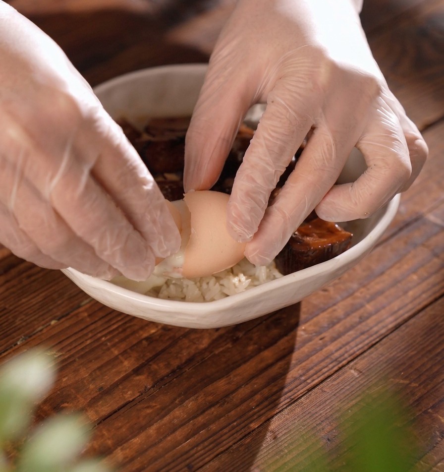 手袋をした手で卵の殻を優しく割り、牛タンが乗った丼の中央に、とろりとした温泉卵を落とし入れている瞬間。