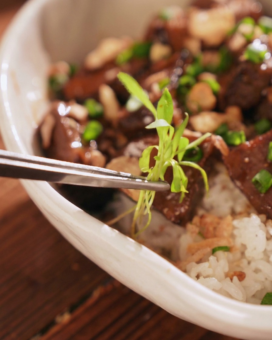 金属製のピンセットを使い、完成した牛タン丼の中央に、繊細な緑色のパクチーの新芽を丁寧に添えている様子。