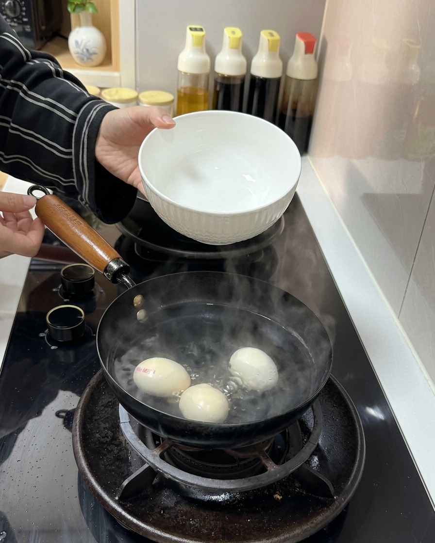 沸騰したお湯と3個の卵が入った鍋に、白い陶器のボウルから冷水を注いでいる様子。