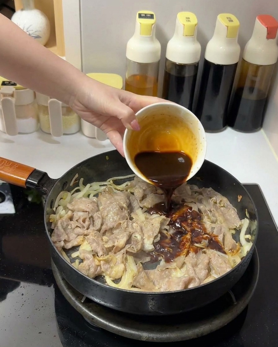 フライパンの中の牛肉と玉ねぎの上に、小さな白い器から濃い茶色のタレを注いでいる様子。