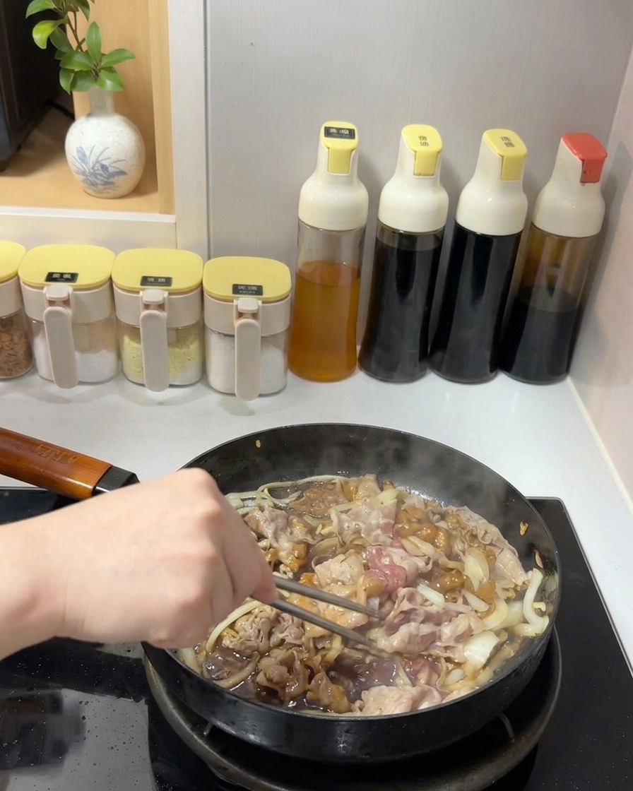 金属製の箸を使って、フライパンの中で牛肉と玉ねぎに濃いツヤのあるタレを絡めながら炒めている様子。