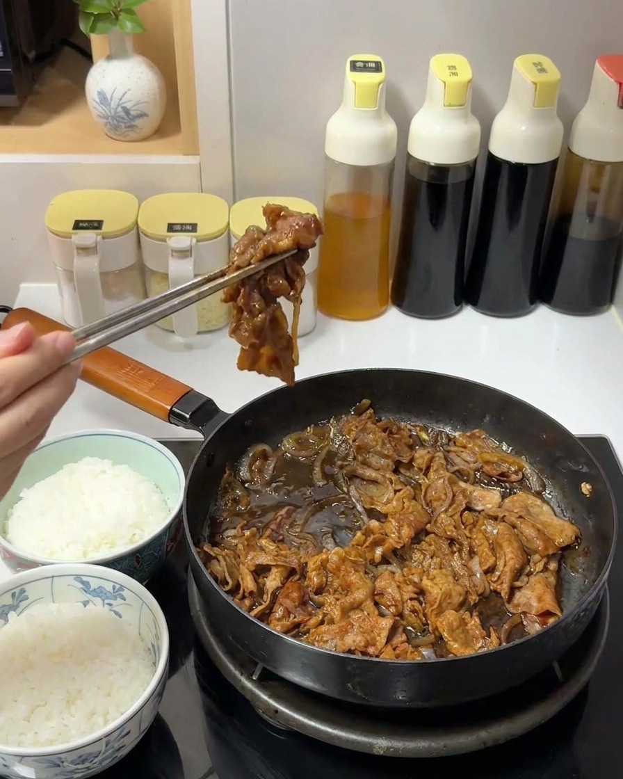 金属製の箸を使い、フライパンからタレがよく絡んだ牛肉と玉ねぎをすくい上げている手。