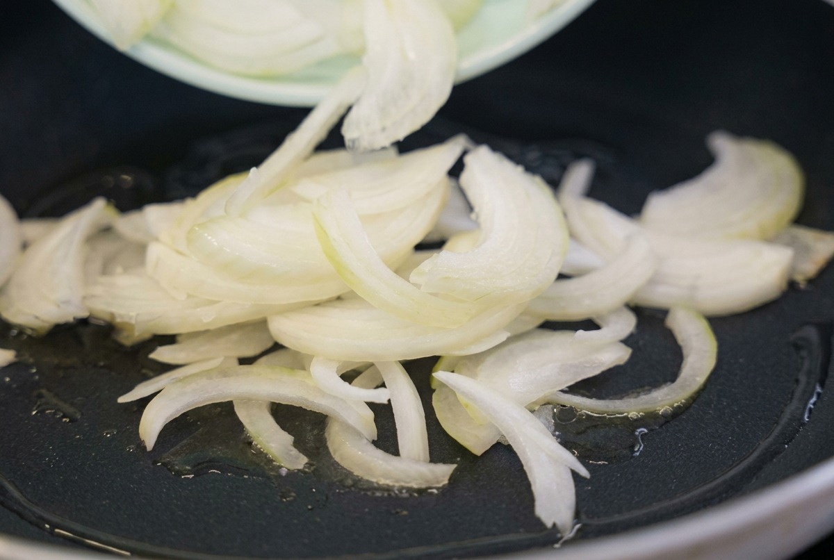 薄切りにした生の白い玉ねぎを、薄緑色のお皿から、油がなじんで温まった黒いフライパンへ流し入れている様子。
